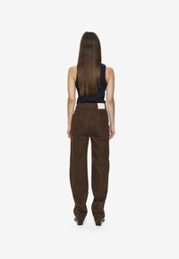 Donna con capelli lunghi e lisci che indossa un top senza maniche nero e pantaloni in stampa pitone marrone, in piedi di spalle contro uno sfondo neutro.