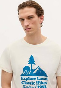 Tricou din bumbac de culoare crem, cu un grafic albastru care prezintă munți, un copac și un soare, având text dedesubt într-un font îndrăzneț.