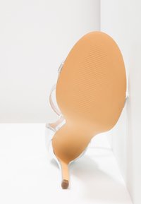 Sandale à talons hauts argentée avec un bout pointu, un talon aiguille mince, des lanières transparentes et une semelle lisse et texturée.