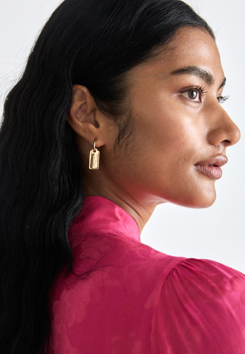 Boucles d'oreilles rectangulaires dorées avec une texture lisse et des détails gravés, portées par un mannequin aux cheveux noirs ondulés, sur un fond clair.