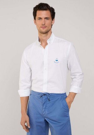 Camisa blanca de botones con logo bordado azul, mangas largas y puños enrollados, combinada con pantalones cortos de tiro medio en azul claro con cordón.