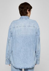Camicia in denim azzurro chiaro con spalla posteriore, maniche lunghe e polsini con bottoni. Il tessuto ha una texture scolorita con dettagli di cuciture sottili.