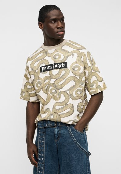 Μπεζ κοντομάνικο μπλουζάκι με σχέδιο φιδιού, με μαύρο λογότυπο "Palm Angels" στο στήθος. Φορώντας με τζιν σορτς.