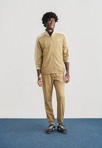 Καφέ κοντή ζακέτα με λευκές πλαϊνές ρίγες, φερμουάρ μπροστά, συνδυασμένη με παντελόνι ίδιου σχεδίου, υφασμάτινη υφή, φορεμένη με μαύρα αθλητικά παπούτσια.