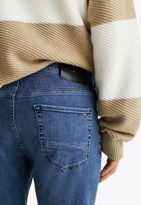 Vaqueros de mezclilla de color azul oscuro con un detalle en el bolsillo trasero y un parche de cuero negro que presenta un logo. Se puede ver un suéter de punto a rayas en tonos beige y crema.