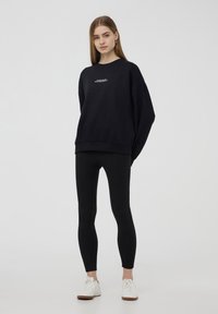 Felpa nera oversize con testo bianco, abbinata a leggings neri e sneakers bianche. Design semplice e casual con una vestibilità comoda.
