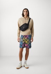 Borsa a tracolla nera con scomparti con cerniera, indossata sopra un felpa beige con cappuccio e pantaloni shorts a fantasia multicolore. Sneakers bianche completano l'outfit.