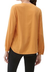 Blouse jaune moutarde avec manches longues bouffantes et col rond. Tissu texturé avec motifs diagonaux, ourlet arrondi. Portée avec un jean noir.