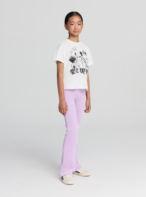 T-shirt bianca corta con design grafico nero, abbinata a pantaloni svasati lilla a coste e scarpe beige, in piedi su uno sfondo semplice.