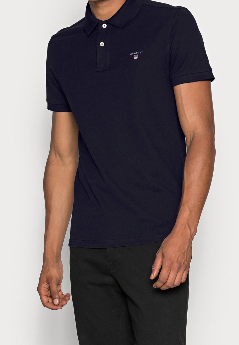 Marinblå polo med klassisk krage, tvåknappsknäppning och broderad logotyp. Tyget är slätt och åtsittande, i kombination med svarta byxor.