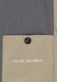 Tessuto grigio scuro con un pannello beige caratterizzato da un bottone nero e testo ricamato bianco "OSCAR_JACOBSON." Superficie testurizzata, design su misura.
