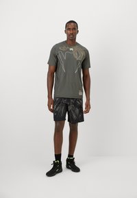 T-shirt de sport à manches courtes en vert olive, avec un design graphique doré ; associé à un short noir affichant un motif audacieux et des baskets noires.