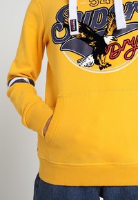 Sudadera amarilla con un gráfico de un águila y el texto "Superdry". Cuenta con un bolsillo frontal, puños acanalados y una capucha con cordón.