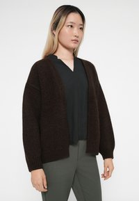 Brun strikket cardigan med løs passform, åpen foran og lange ermer, brukt over en svart topp og kombinert med grønne bukser.