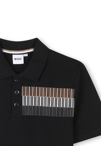 Black polo shirt featuring a horizontal piano key design in brown, white, and black. Four-button placket and textured fabric. Brand label visible.