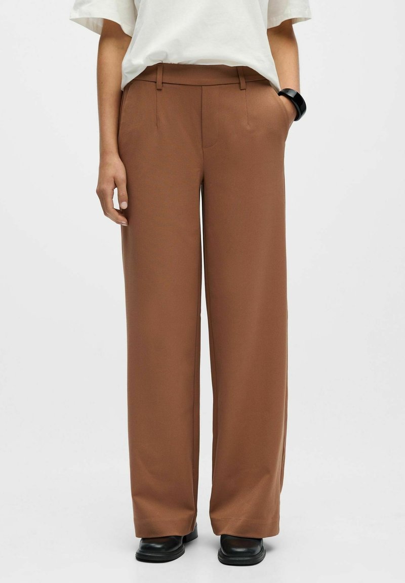 Personne portant un pantalon marron à taille haute et larges jambes, une chemise blanche à manches courtes, des chaussures noires et un bracelet noir, debout devant un fond uni.