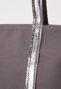 Pilka audinio pirkinių maišelį su blizgučiais sidabriniu ruožu ant rankenos, pabrėžiančiu jo tekstūrą ir dizaino ypatybes.