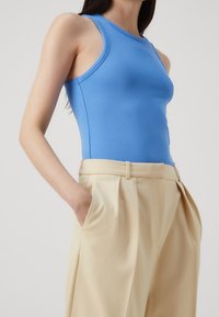 Blaues geripptes Tanktop, kombiniert mit hochgeschnittenen, gefalteten beigen Hosen. Der Stoff wirkt glatt und hat eine figurbetonende Silhouette.