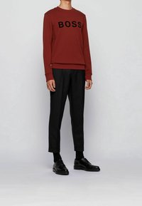 Sweat-shirt rouge avec le logo "BOSS", associé à un pantalon de costume noir. Des chaussures noires complètent la tenue. Tissu lisse, coupe ajustée.