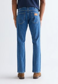 Oseba z modrimi kavbojkami z ravnimi hlačnicami in rjavimi škornji iz semiša, stoji pred enobarvnim svetlim ozadjem, vidno od zadaj.