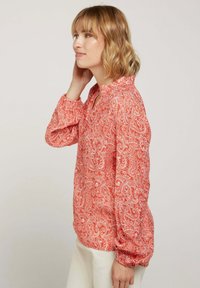 Blouse rouge à motif paisley avec de longues manches bouffantes et un décolleté en V. Tissu léger avec une texture douce et une coupe décontractée.