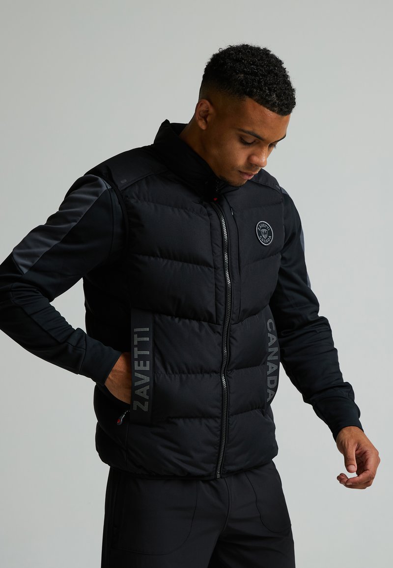 Gilet rembourré noir avec fermeture éclair, présentant un logo sur la poitrine et un texte réfléchissant sur le côté. Fabriqué en tissu lisse avec une coupe ajustée.