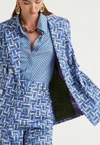 Costume bleu à motifs avec un design géométrique sur le blazer et le pantalon. La chemise en dessous présente un petit motif bleu. La doublure est violette.