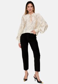 Weiße Bluse mit Ballonärmeln, floraler Umrissdruck und Schleifenbund, kombiniert mit schwarzen, eng geschnittenen Cordhosen und schwarzen spitzen High Heels.