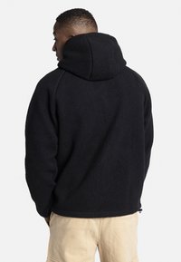 Hoodie en polaire noir avec une surface texturée, grande capuche et coupe décontractée, associé à un pantalon de couleur claire. Présente des zips latéraux.