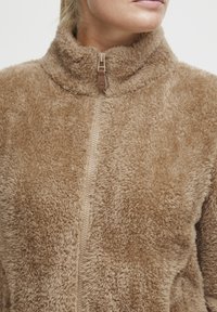 Tan fleece jacket with a high collar, zipper front, and plush texture. Features a smooth zipper and a soft, shaggy appearance.