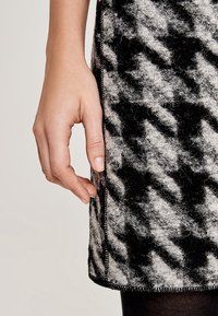 La main gauche repose contre une jupe en laine à motifs noir et blanc avec une couture latérale visible et un ourlet détaillé.