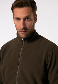 Giacca in pile marrone con colletto alto e zip frontale. Il tessuto ha una consistenza morbida, offrendo calore e comfort.