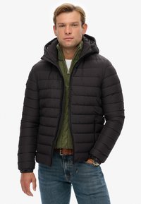 Sort puffjakke med lynlås foran og hætte, der har kviltede sektioner, båret over en grøn kabelstrikket sweater, parret med blå jeans.
