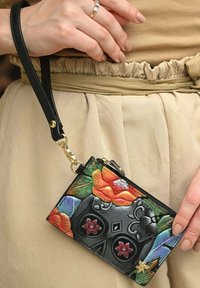 Eine Hand hält eine kleine schwarze Handgelenktasche mit buntem, geprägtem Blumen- und Totenkopfdesign, die mit beigen, gerafften Bundhosen getragen wird.