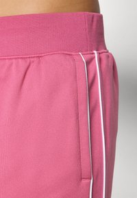 Roze trenirke s rebrastim pojasom, bočnom džepom i bijelim detaljima uz šav. Tkanina izgleda mekano i rastezljivo.