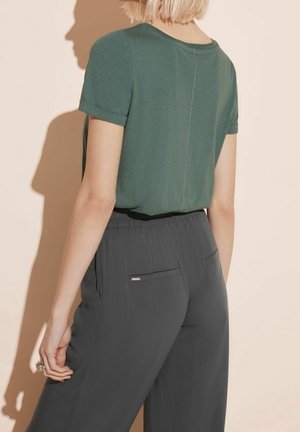 Donna con capelli biondi corti che indossa una maglietta verde a maniche corte infilata in pantaloni grigio scuro con vita elastica, vista da dietro.