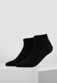 Ein Paar schlichte schwarze Knöchelsocken auf einem weißen Podest vor einem hellgrauen Hintergrund präsentiert.