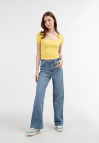 Tricou galben cu manșete scurte și decolteu pătrat, cu bretele cruzate, asortat cu blugi largi de culoare albastră deschisă și adidași multicolori.