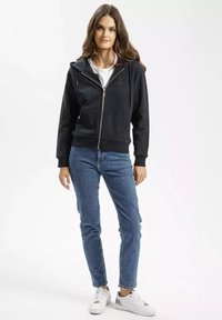 Czarna bluza z zamkiem i kieszenią z przodu, noszona na białej koszulce. W zestawie z niebieskimi dżinsami i białymi trampkami. Model stoi na jednolitym tle.