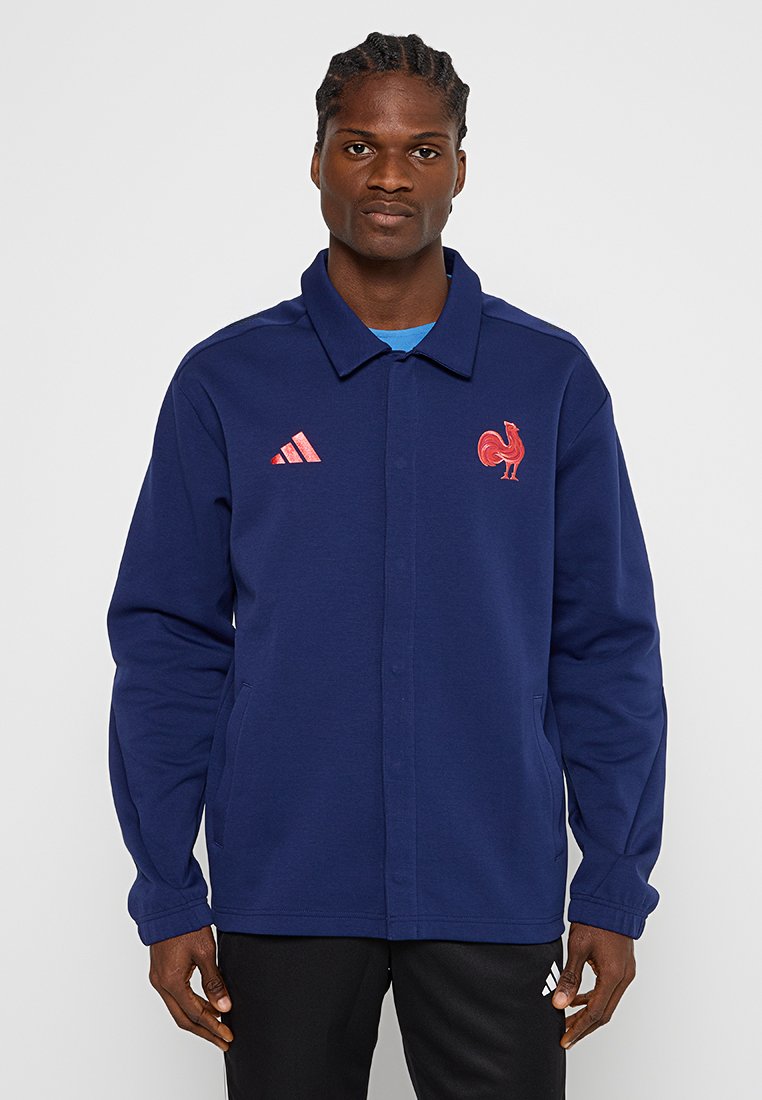 Navyblaues sportliches Jackett mit Knopfverschluss, Kragen und seitlichen Taschen. Es verfügt über ein rotes Hühnerlogo und drei gestreifte Akzente an den Ärmeln.