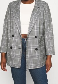 Rutig dubbelknäppt blazer i grå och svarta nyanser, med slitsade kragar och två framfickor, över en vit topp och blå jeans.