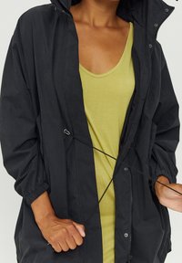 Czarna lekka kurtka z troczkiem, długimi rękawami i luźnym krojem, noszona na jasnzie zielonym topie na ramiączkach. Gładka faktura, casualowy design.