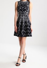 Marinblå ärmlös klänning med blommönster i vitt, utsvängd kjol och åtsittande byst. Bärs med svarta klacksandaler med silverdetaljer.