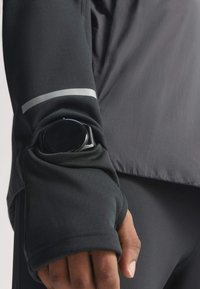 Black long-sleeve jacket with thumb hole sleeve pocket holding a black smartwatch on a person’s wrist.