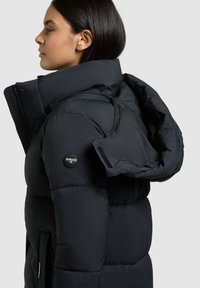 Zwarte pufferjas met hoge kraag, ritssluiting en een grote capuchon. Heeft een logopatch op de linkermouw en een gewatteerde textuur.