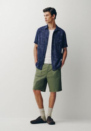 Junger Mann steht und blickt nach rechts, trägt ein marineblaues kurzärmeliges Hemd über einem weißen T-Shirt, grüne Cargoshorts, beige Socken und braune Slipper.