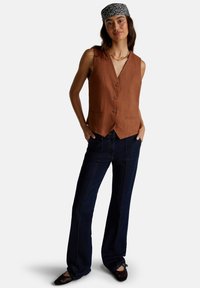 Gilet in lino marrone con cinque bottoni, indossato su jeans svasati blu scuro. Modello accessorato con un cerchietto nero a fantasia e scarpe basse.