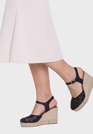 Nogi w czarnych perforowanych sandałach na koturnie z paskami wokół kostki i plecionym sznurowym obcasem, w połączeniu z jasnoróżową spódnicą do kolan.