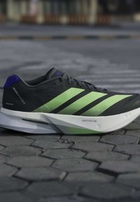 Μαύρο και πράσινο παπούτσι για τρέξιμο Adidas Adizero Boston 13 με λευκή σόλα Lightstrike Pro πάνω σε πλακόστρωτο έδαφος.