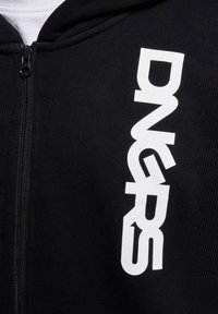 Haut à fermeture éclair noir en tissu côtelé, arborant un grand logo blanc "DNGRS" imprimé verticalement sur le côté droit.